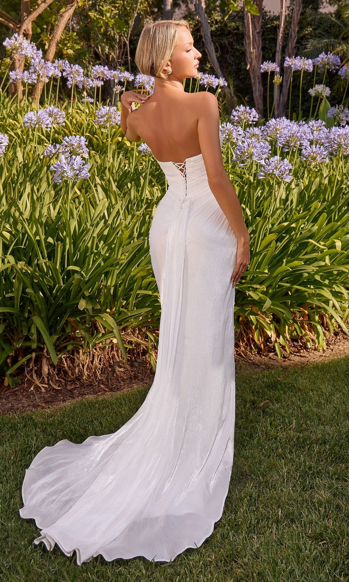 A woman in the Ladivine CD411W long formal dress stands outdoors on grass, facing away, surrounded by green plants and purple flowers.